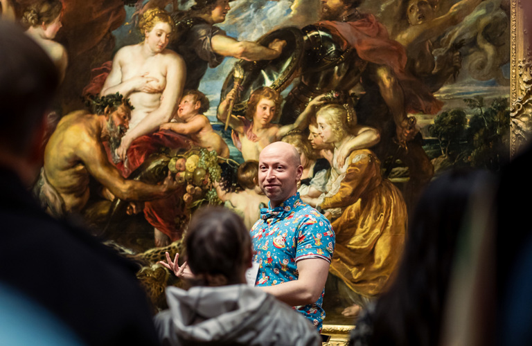 A photograph of a man stood in front of a large painting that seems to depict feasting and war. He is wearing a bright blue patterned shirt and is giving a talk to a crowd of people.