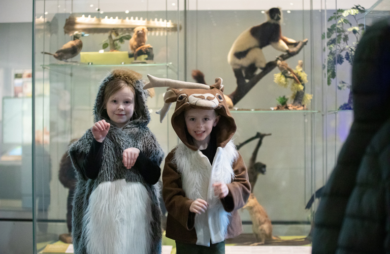A photograph of two children dressed in animal dress up clothing, photographed in front of a taxidermy monkey. 