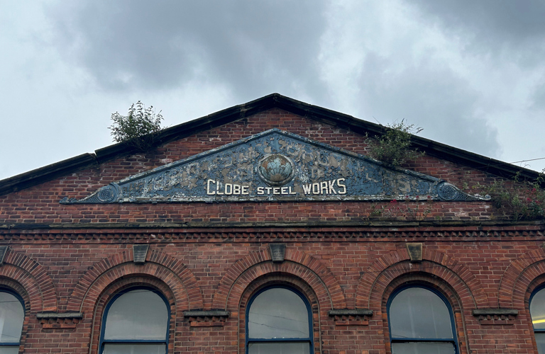 A photo of the top of an old red brick industrial building. at the bottom of the image is a row of windows which are curved at the top. At the top of tech building is a triangular sign that says Globe Steel Works.