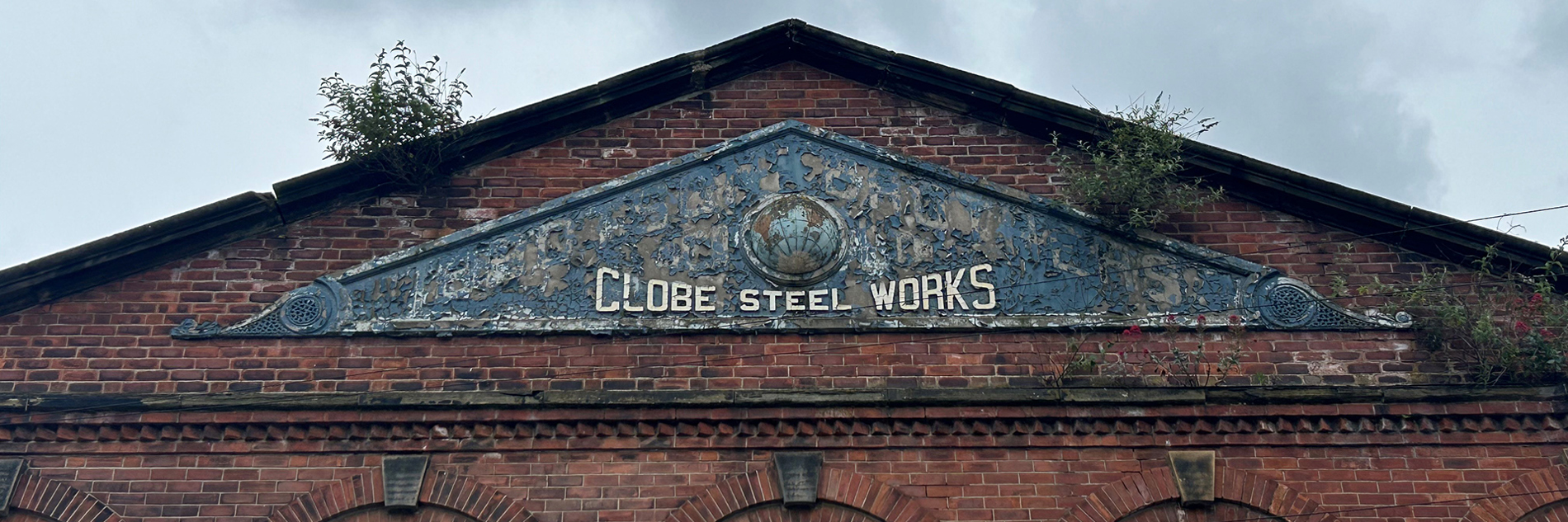 A photo of the top of an old red brick industrial building. at the bottom of the image is a row of windows which are curved at the top. At the top of tech building is a triangular sign that says Globe Steel Works.