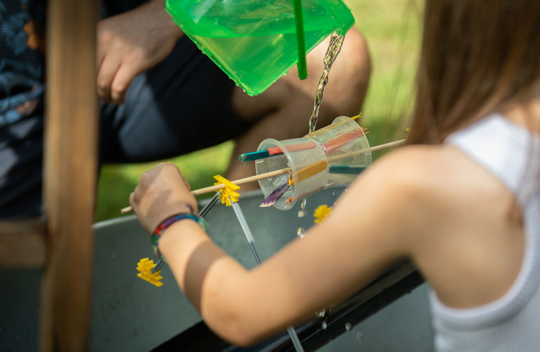 A child testing a crafted waterwheel, made from plastic cups and lollipop sticks.