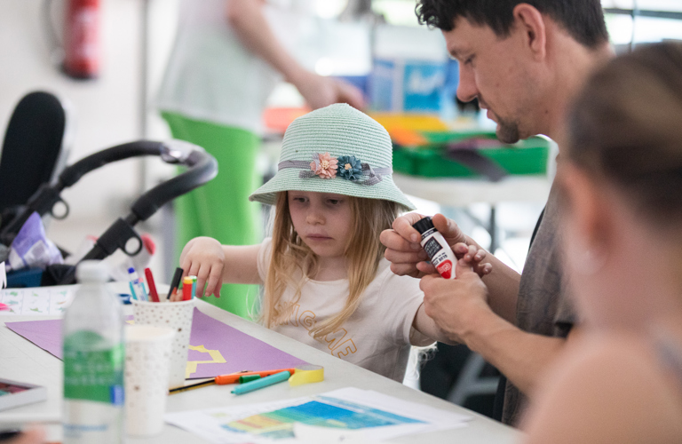 A photograph of a young white child being assisted in a crafting activity by an older white male. The child wears a floral hat and there is lots of paper on the table.