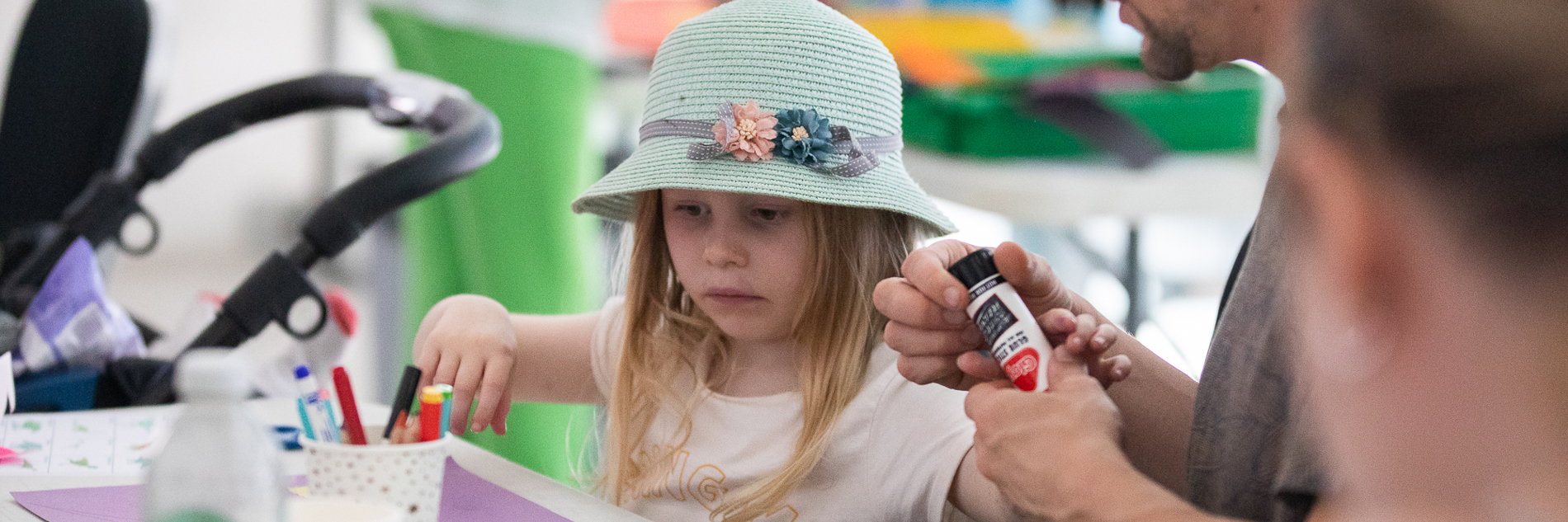 A photograph of a young white child being assisted in a crafting activity by an older white male. The child wears a floral hat and there is lots of paper on the table.
