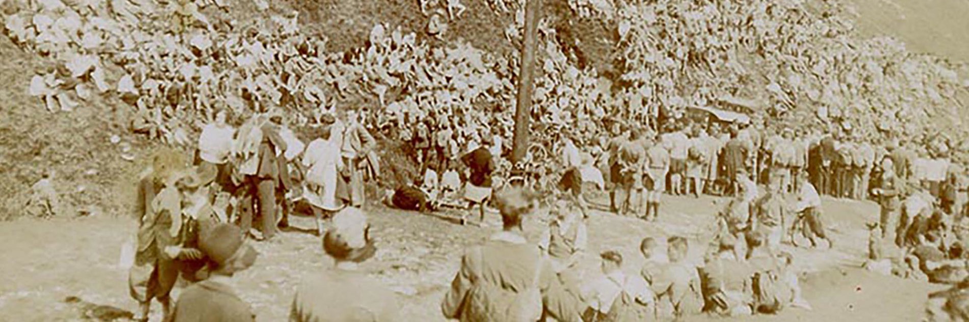 A sepia monochrome photo of a large crowd of adults and young people who are sat and stood on the hillside next to a large wooden pole.