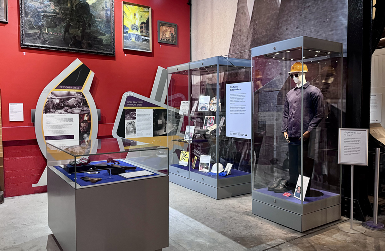 A photograph of displays in a museum including large paintings of industrial scenes and cases with objects including a steelworks hardhat and workwear, tools, and a decorative plate. 