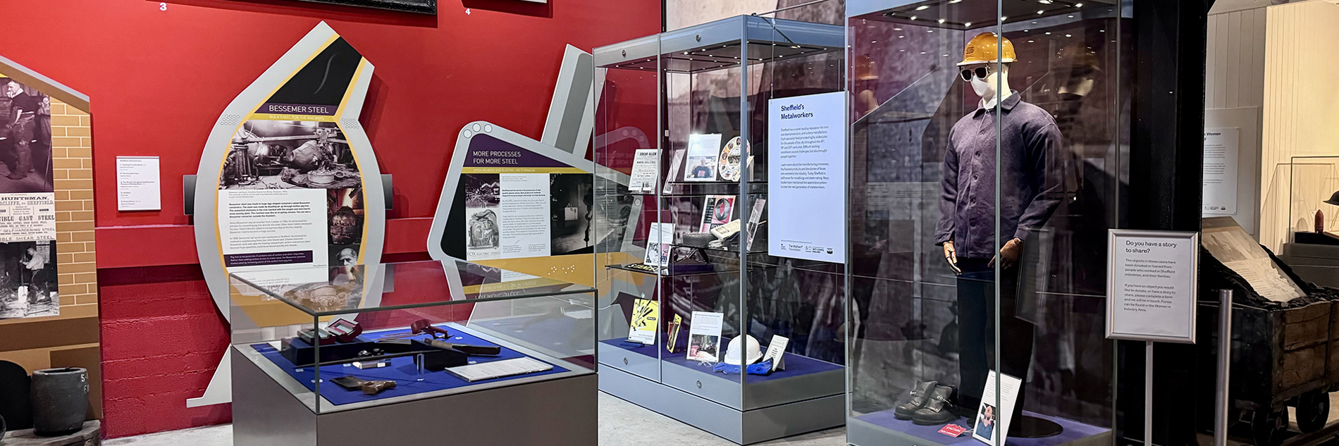 A photograph of displays in a museum including large paintings of industrial scenes and cases with objects including a steelworks hardhat and workwear, tools, and a decorative plate. 