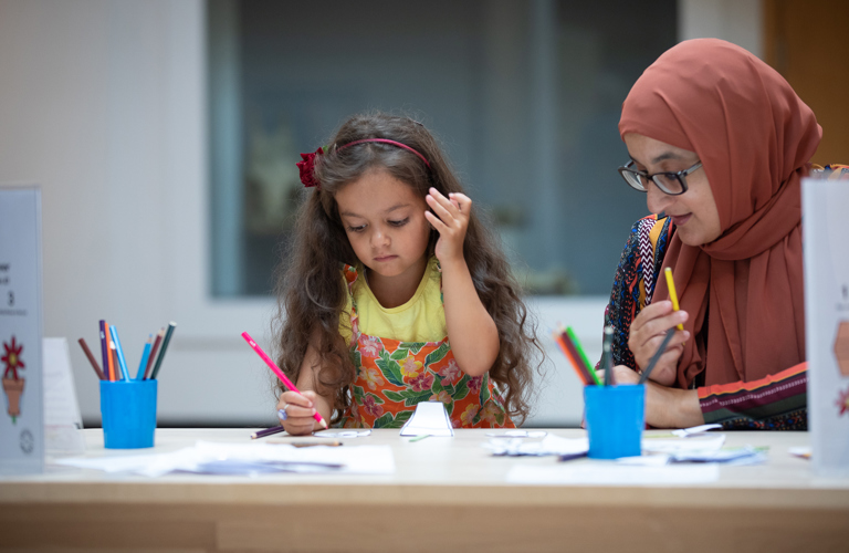 A young girl and her mother sit at a table drawing, with two blue pots of pencils visible on the table in front of them
