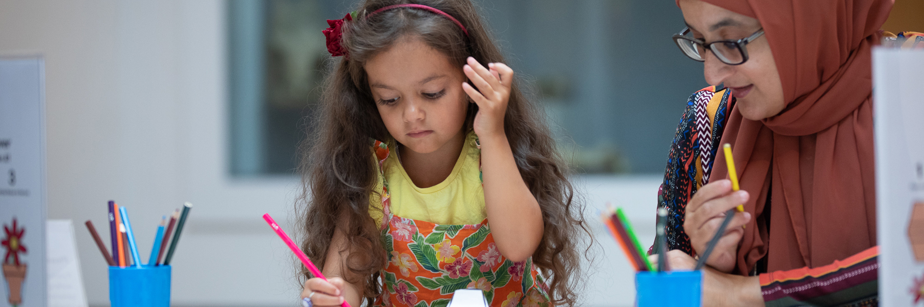 A young girl and her mother sit at a table drawing, with two blue pots of pencils visible on the table in front of them