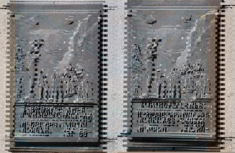 A photograph of a grey metal plaque on a sandy cream brick wall. The plaque reads ' To commemorate all ramblers who campaigned for national parks and public access to mountains and moorland. Year 2000'. Above the text, there are engravings showing groups of people, most of whom are wearing backpacks, exploring a hilly countryside.