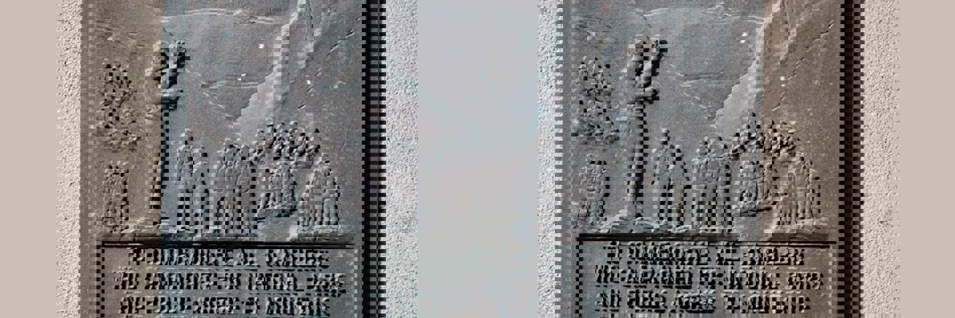 A photograph of a grey metal plaque on a sandy cream brick wall. The plaque reads ' To commemorate all ramblers who campaigned for national parks and public access to mountains and moorland. Year 2000'. Above the text, there are engravings showing groups of people, most of whom are wearing backpacks, exploring a hilly countryside.