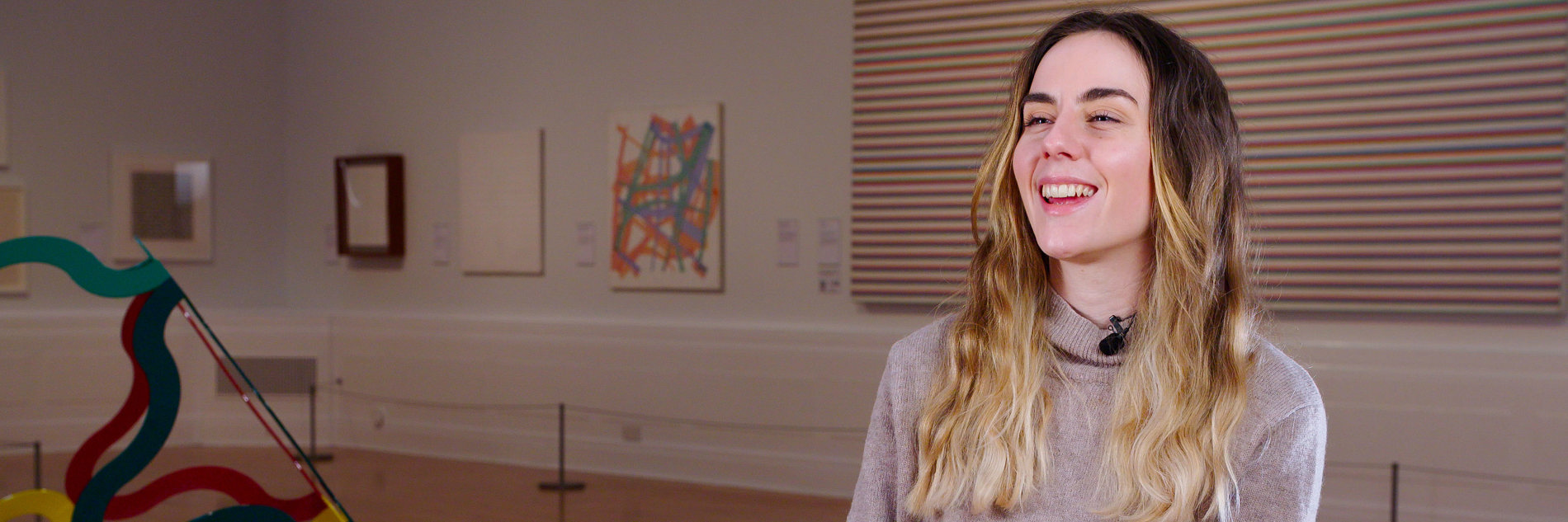 A photograph of an adult sitting on a chair in an art gallery. In the front of them is a sculpture and behind them are several artworks on the wall with a small barrier in front of them. 