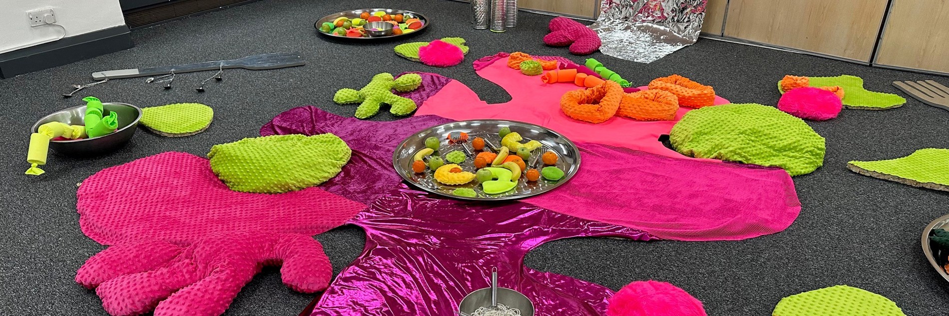 A photograph of an indoor space set up for toddler play, with lots of soft cushions, shiny fabric, and sensory toys.
