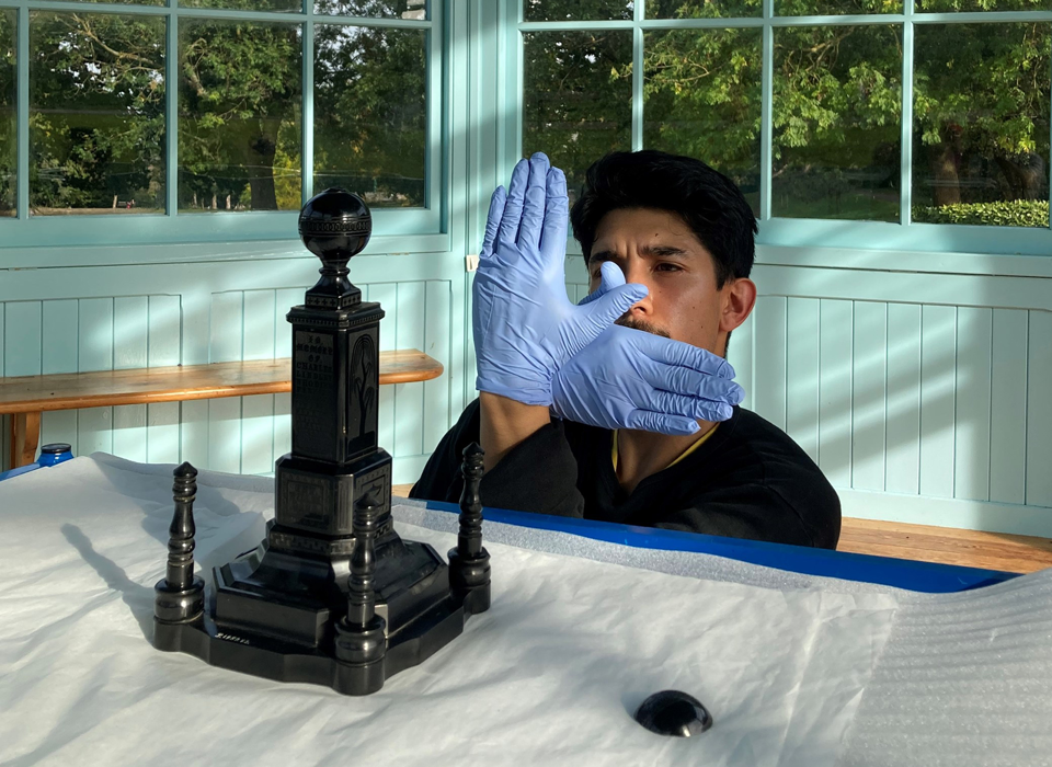 A person behind a table wearing blue gloves making an overlapping hand gesture in an L shape. On the table is a small version of a monument, with four columns surrounding a large central square tiered column topped with a sphere.