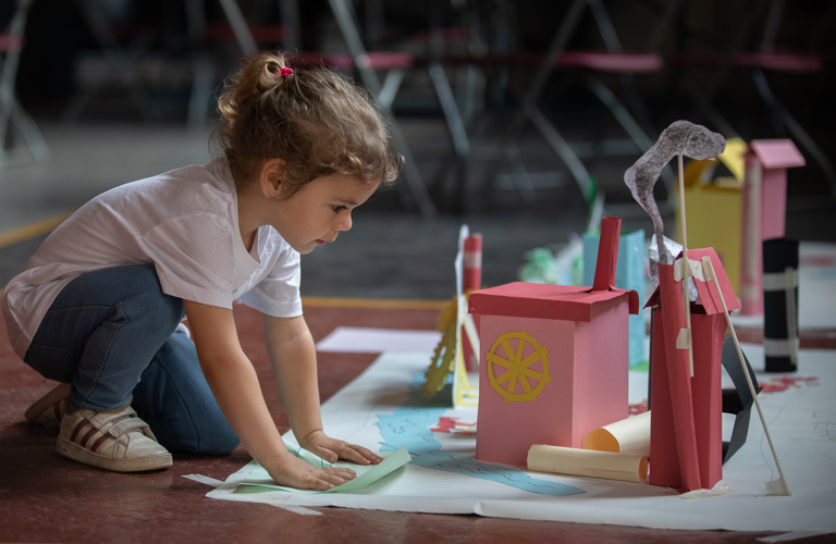 A photograph of a child with brown hair crouching in front of a display of hand crafted paper houses, with wheels and smoke also added in paper.