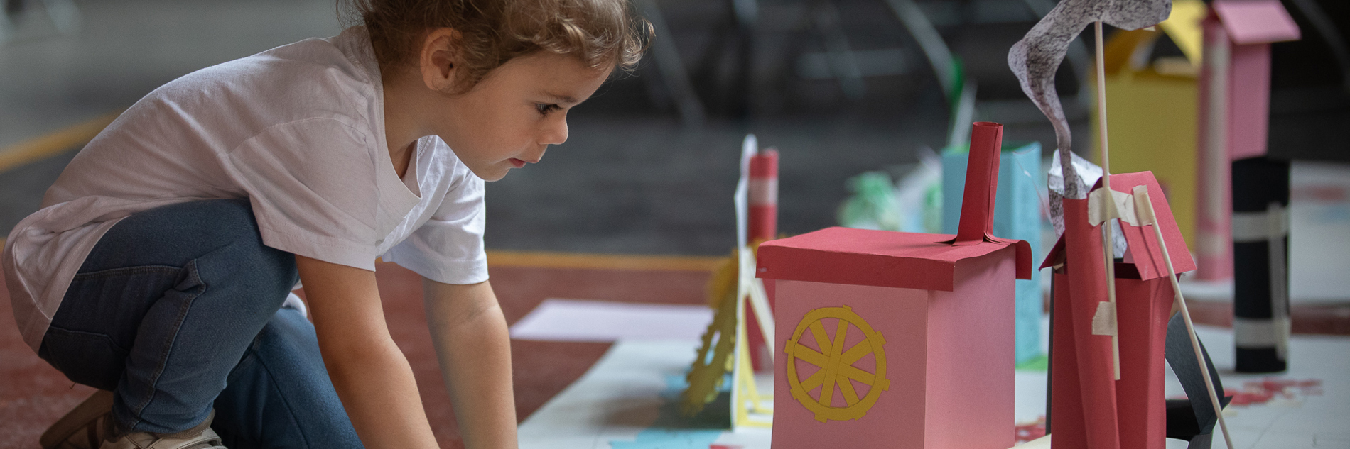 A photograph of a child with brown hair crouching in front of a display of hand crafted paper houses, with wheels and smoke also added in paper.