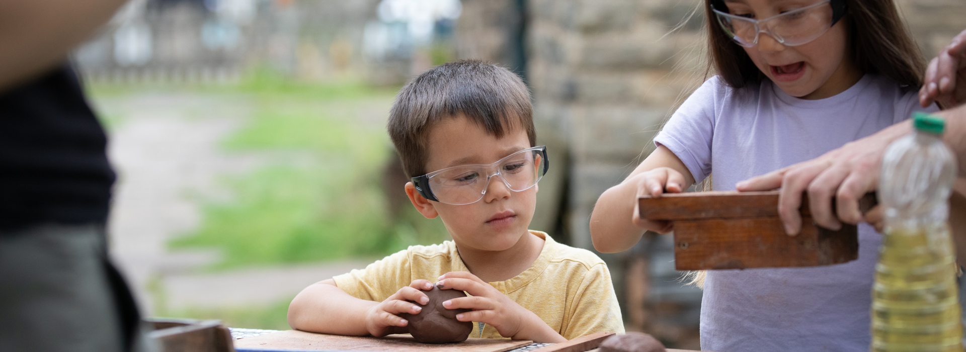 A photograph of two children with brown hair wearing goggles and rolling brown clay. One holds a wooden mold.