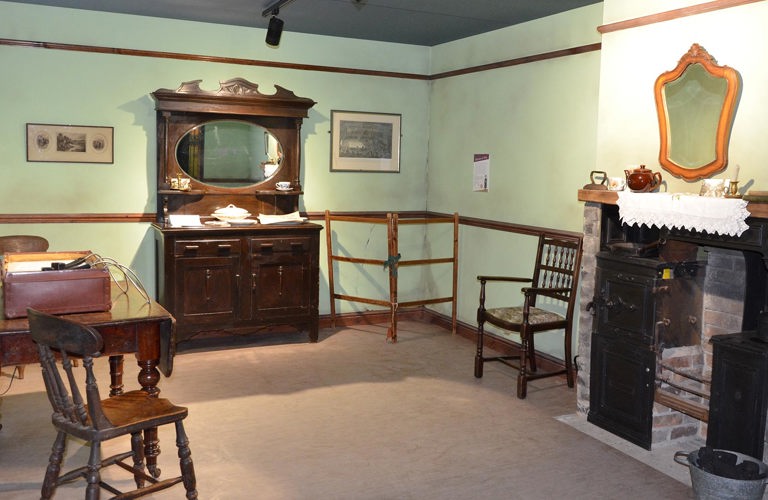 A living room with pale green walls containing items of dark oak furniture including a dining table and chairs and a large ornate mirrored dresser. There is a clothes drying frame in the corner and part of an old Yorkshire Range fireplace is visible to one side.