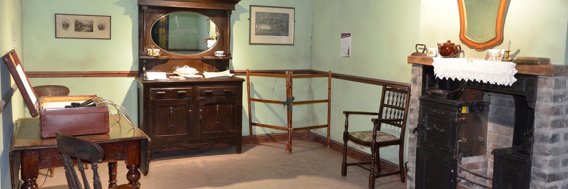 A living room with pale green walls containing items of dark oak furniture including a dining table and chairs and a large ornate mirrored dresser. There is a clothes drying frame in the corner and part of an old Yorkshire Range fireplace is visible to one side.