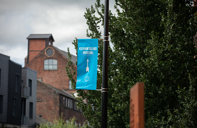 A photograph of a blue banner that reads 'Kelham Island Museum' in white text, with a white arrow point towards the museum. There are trees and red brick buildings in the background. The banner is attached to a black lamppost.