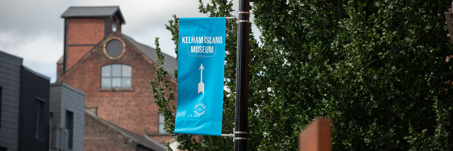 A photograph of a blue banner that reads 'Kelham Island Museum' in white text, with a white arrow point towards the museum. There are trees and red brick buildings in the background. The banner is attached to a black lamppost.