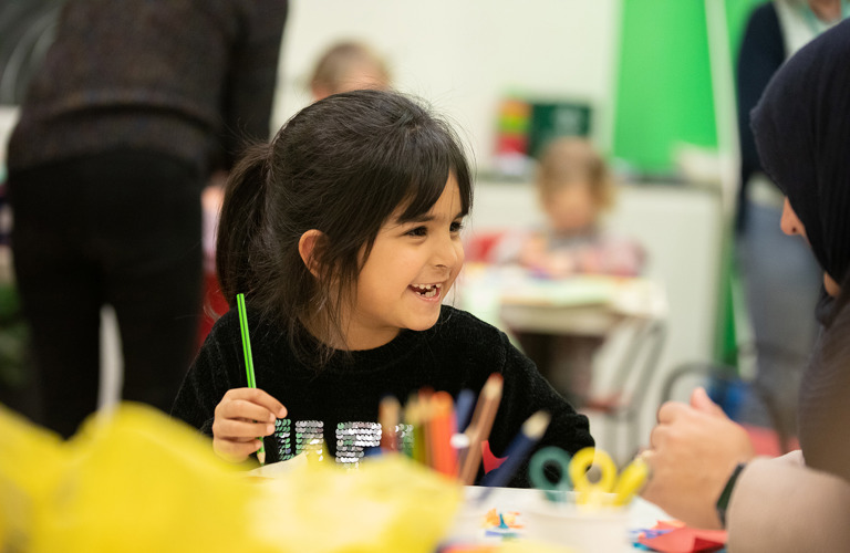 A young child is sitting at a table and is smiling and holding a green pencil. In the foreground is some yellow tissue paper and pots of scissors and pencils.