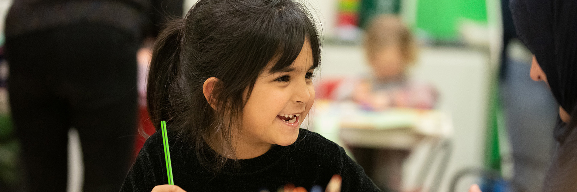 A young child is sitting at a table and is smiling and holding a green pencil. In the foreground is some yellow tissue paper and pots of scissors and pencils.