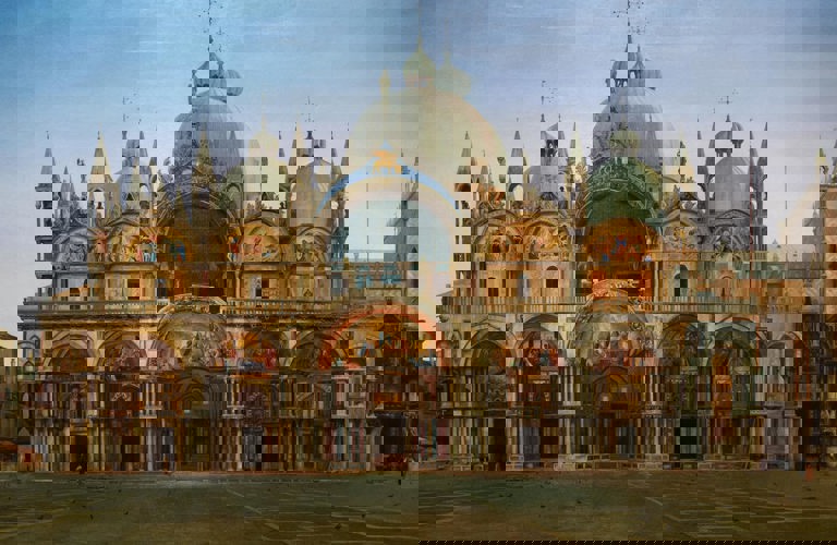 J.W.Bunney's painting Western Façade of the Basilica of San Marco, Venice. The basilica has many impressive golden arches and white domes, inspired by Islamic architecture. The square stretches out in front of, with only very few people depicted. The sky is blue at the top, but looks close to sunrise or sunset. 