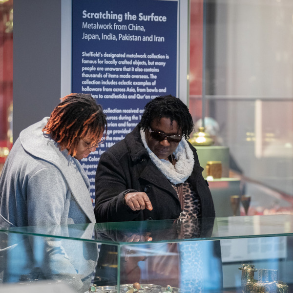 Two adults are looking at a glass cabinet in the gallery, one of the adults is pointing. There is a dark blue gallery sign behind them. 