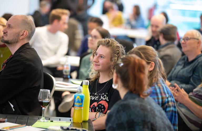 A photograph showing attendees at an event in Millennium Gallery. The room is full of people sat at tables, but the camera focuses on a person at the front with long curly blonde hair. On the table in front of them there is a glass or red wine, a bottle of lucozade and a bright yellow water bottle. There are also pencils, pens and paper on the table. Other people sit at the table and they seem to be in discussion