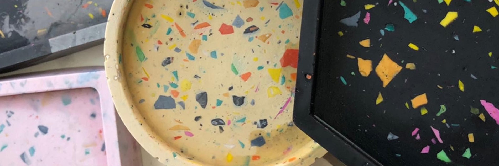 A photograph showing five jesmonite coasters.  There is a square pink one, a square grey one, round white one, a round yellow one and a hexagonal black one. All are speckled with other colours.