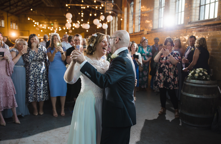 A smiling bride in a wedding dress and a groom in a suit dance in front of wedding guests in a large room decorated with fairly lights and lanterns.