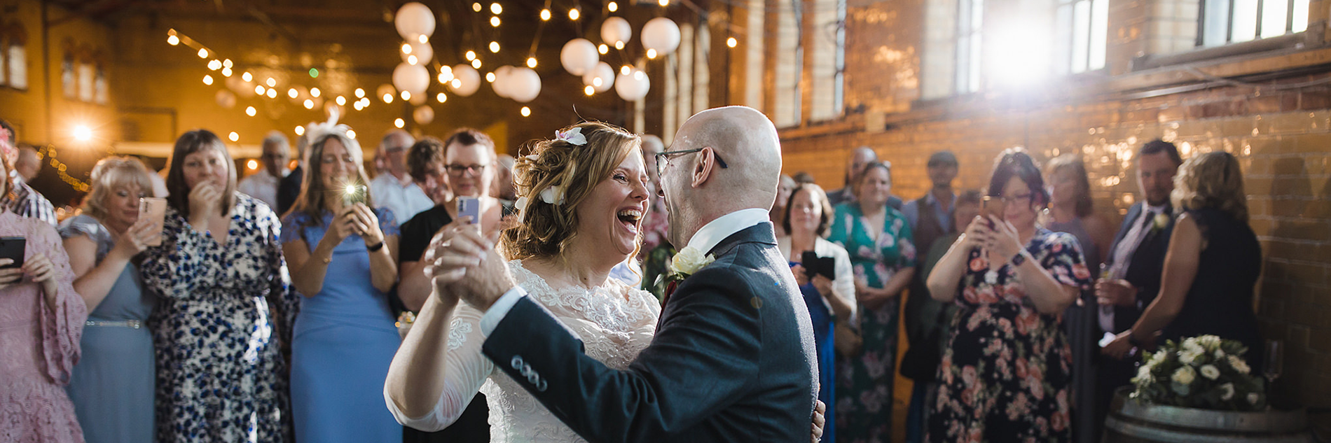 A smiling bride in a wedding dress and a groom in a suit dance in front of wedding guests in a large room decorated with fairly lights and lanterns.