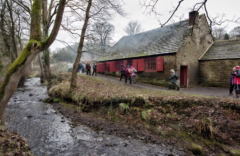 A group of adults walking along a path between a river and a low stone building. There are a number of trees on the banks of the river and a wood in the distance. 