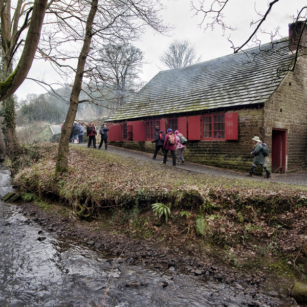 A group of adults walking along a path between a river and a low stone building. There are a number of trees on the banks of the river and a wood in the distance. 