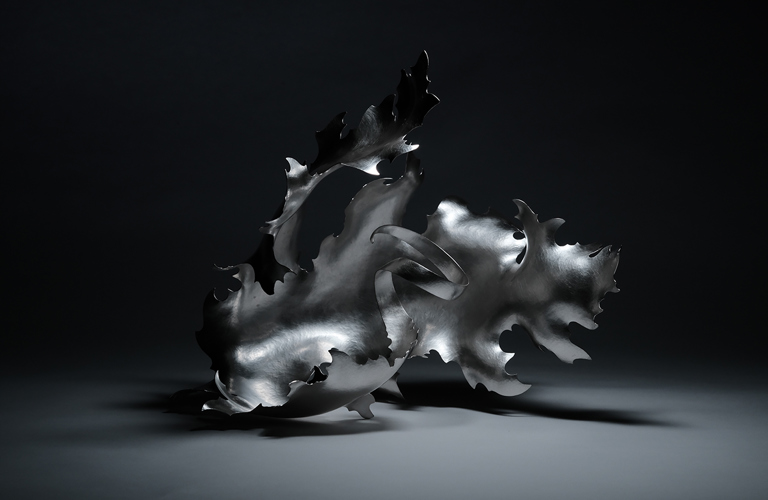 A sculptural, silver coloured metal form reminiscent of a flame, a wave or an exotic plant. It is shown against a monochrome background with high contrast lighting.