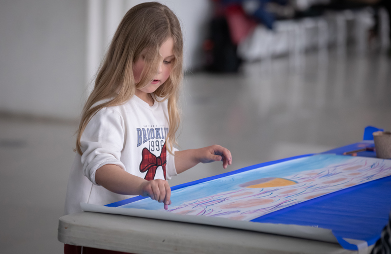 A photograph showing a young girl with long blonde hair painting a sky scene on a long piece of paper. She is wearing a white jumper with a red bow in the centre and is focused on her drawing