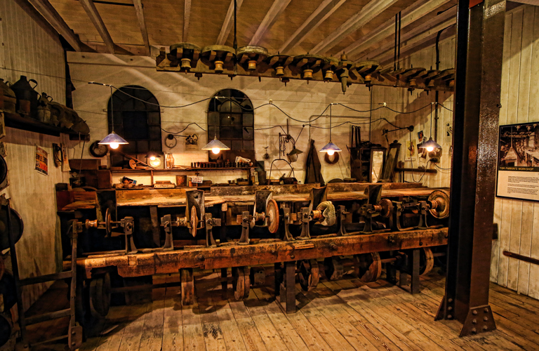 A high and well worn red brick wall fills most of the image. At ground level there are several furnace openings, now bricked up. Old tongs lean against the wall and there are several display boards including a large photograph of the workshop in its working heyday.