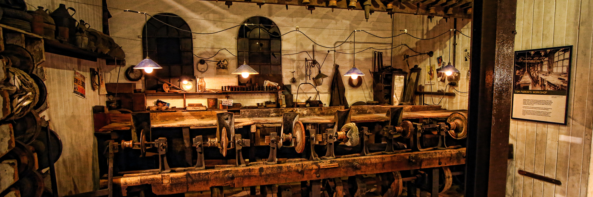 A high and well worn red brick wall fills most of the image. At ground level there are several furnace openings, now bricked up. Old tongs lean against the wall and there are several display boards including a large photograph of the workshop in its working heyday.