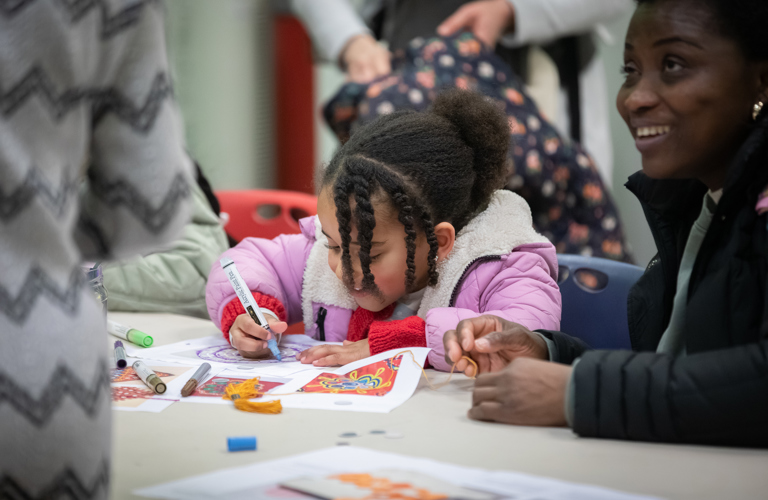 A photograph of a young girl drawing while a woman smiles in conversation with someone next to her. The little girl is focused on the colourful drawing in front of her. There are more pens on the table waiting to be used.