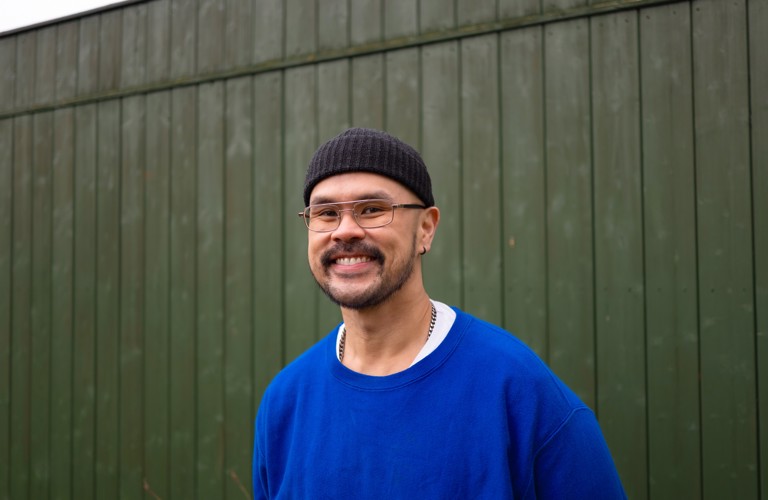 A photograph of a man in a blue jumper, black beanie hat and wire-framed glasses smiling widely at the camera. 