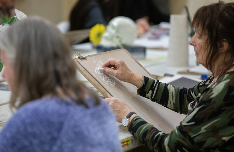 Two adults sitting at a large table. One adult is drawing on an easel. There are art supplies on the table and other adults can be seen in the background. 