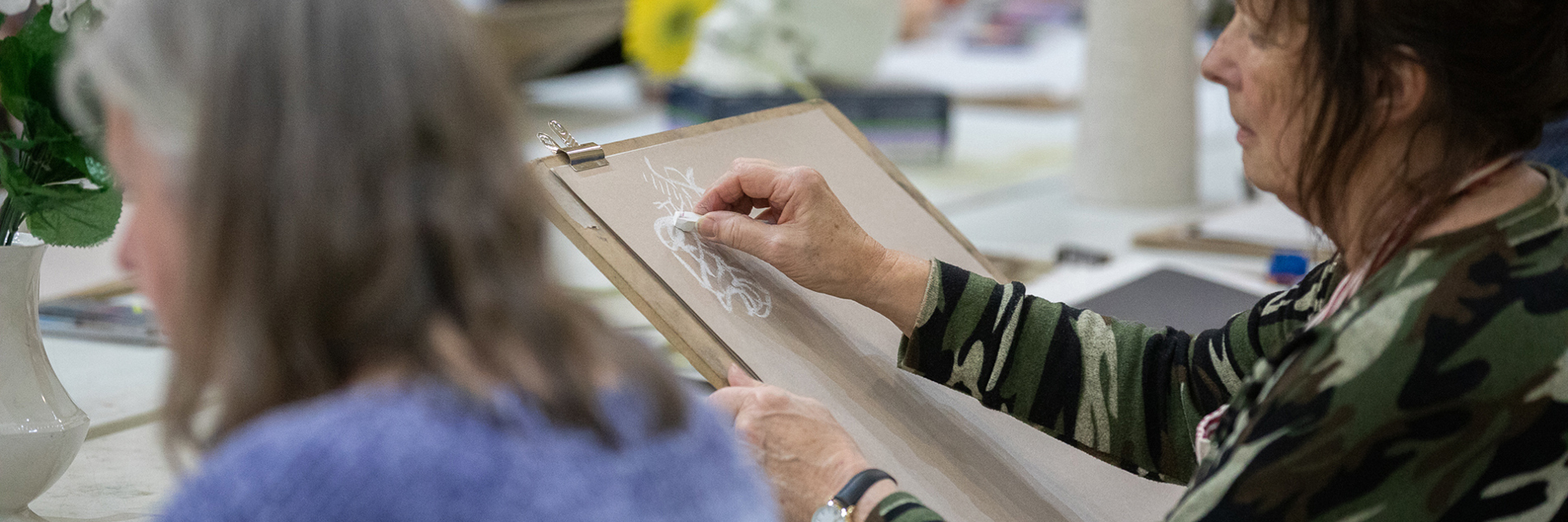 Two adults sitting at a large table. One adult is drawing on an easel. There are art supplies on the table and other adults can be seen in the background. 