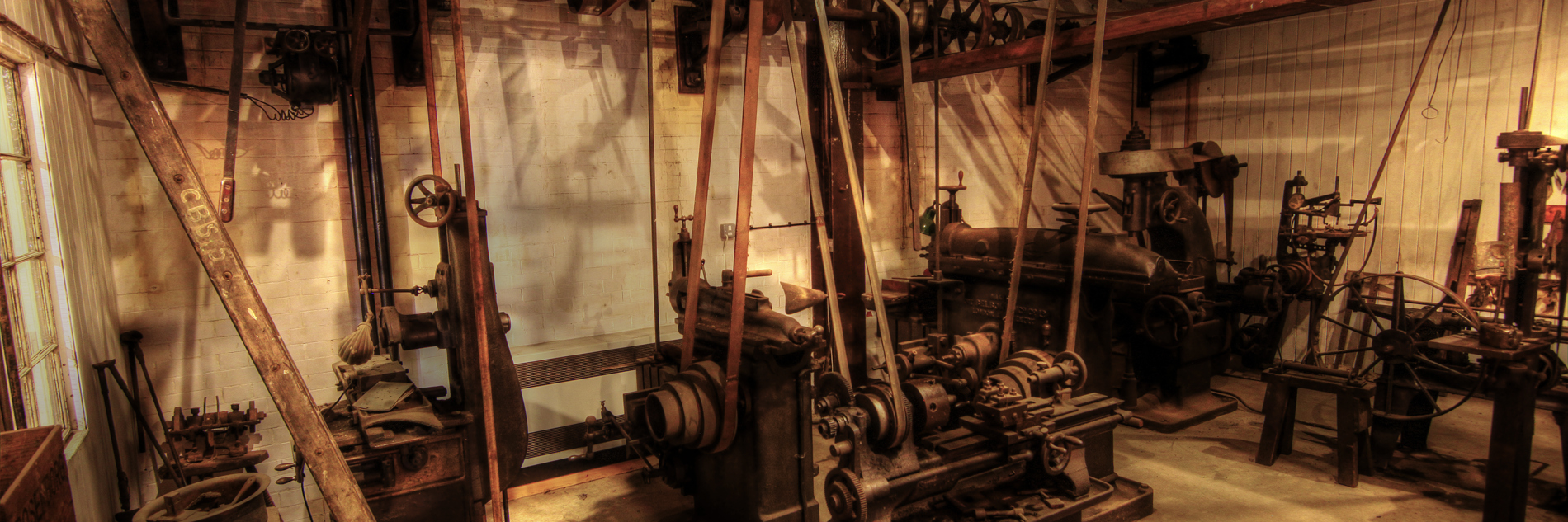 A dimly lit belt driven workshop with several freestanding machines, including a lathe, shaper and milling machine. The drive belts connected to a system of line shafts near the ceiling are a prominent feature.