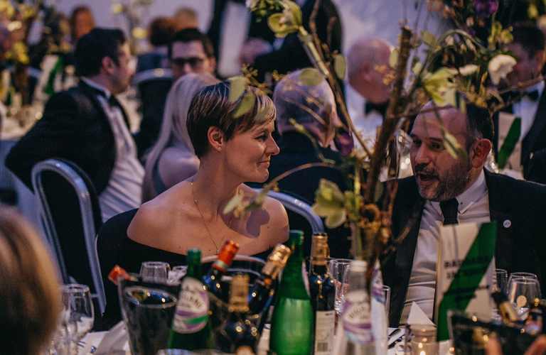 Two formally dressed adults seated at a table. In the background there is another group of seated formally dressed adults. 