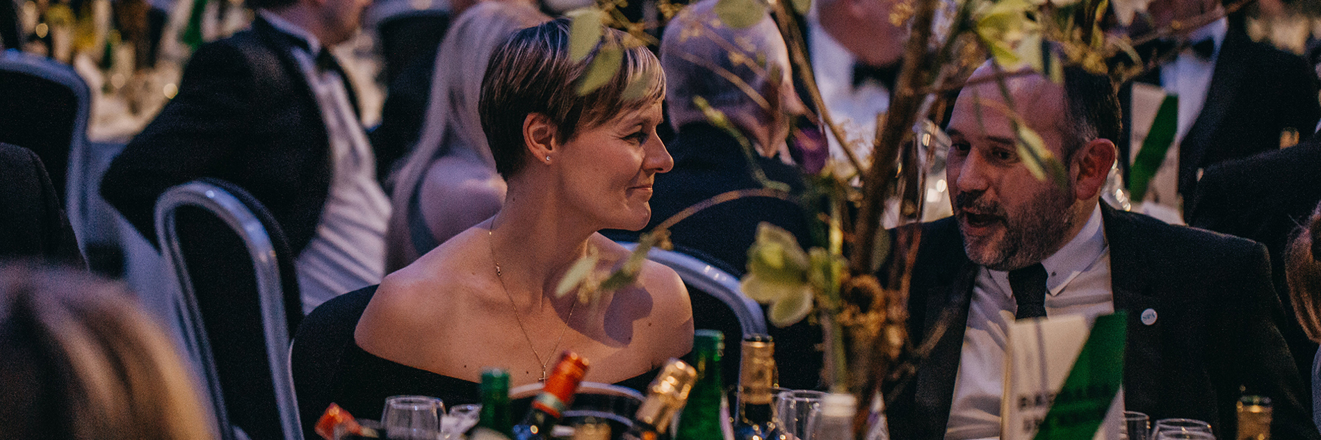 Two formally dressed adults seated at a table. In the background there is another group of seated formally dressed adults. 