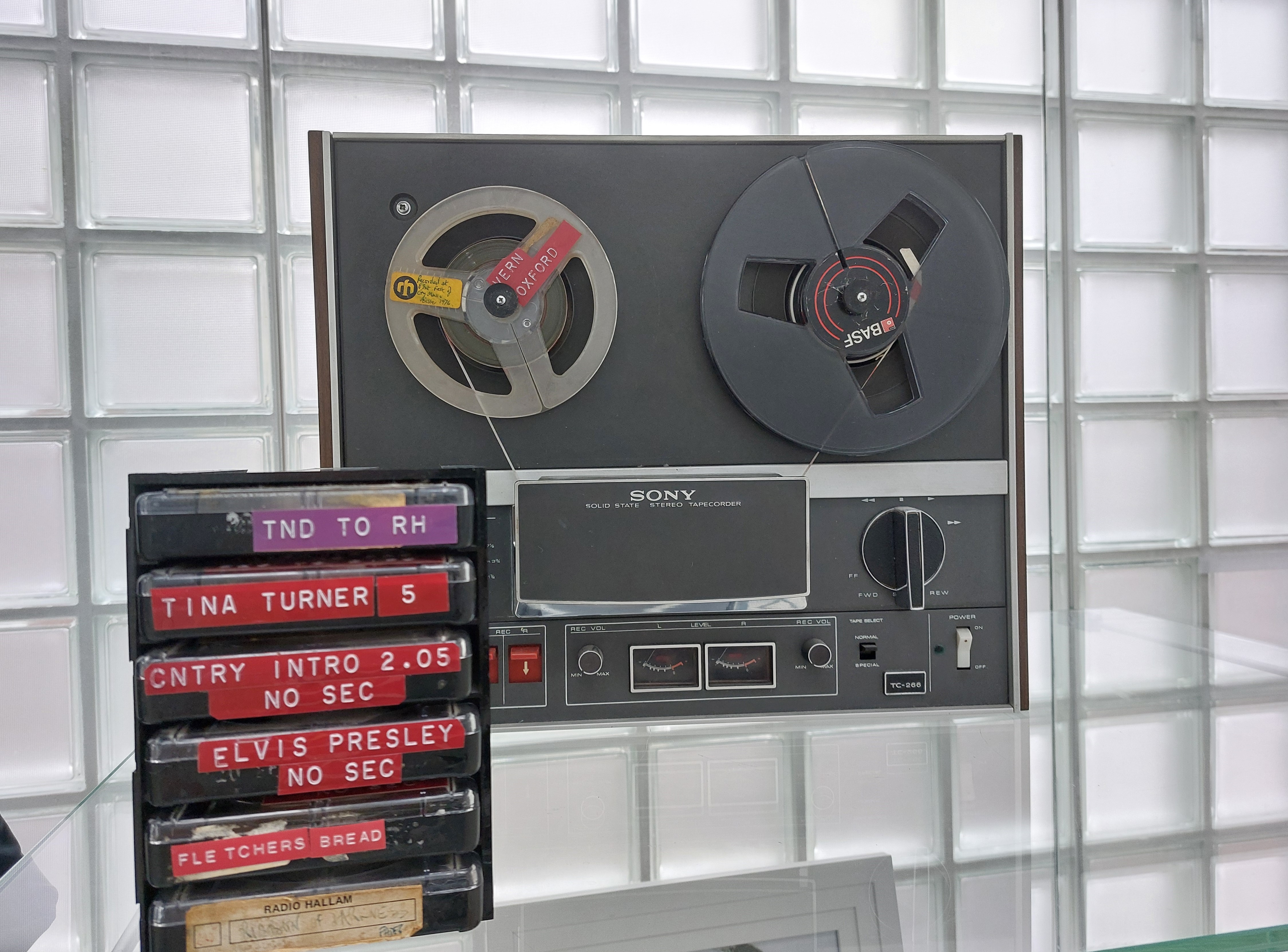 A photograph of radio equipment on display in Millennium Gallery. There is a large machine with two reels in, then a stack of cassette tapes. They are labelled from top to bottom, as follows: TND TO RH, TINA TURNER 5, CNTRY INTRO 2.05 NO SEC, ELVIS PRESLEY NO SEC, and FLETCHERS BREAD