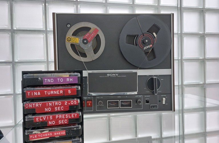 A photograph of radio equipment on display in Millennium Gallery. There is a large machine with two reels in, then a stack of cassette tapes. They are labelled from top to bottom, as follows: TND TO RH, TINA TURNER 5, CNTRY INTRO 2.05 NO SEC, ELVIS PRESLEY NO SEC, and FLETCHERS BREAD