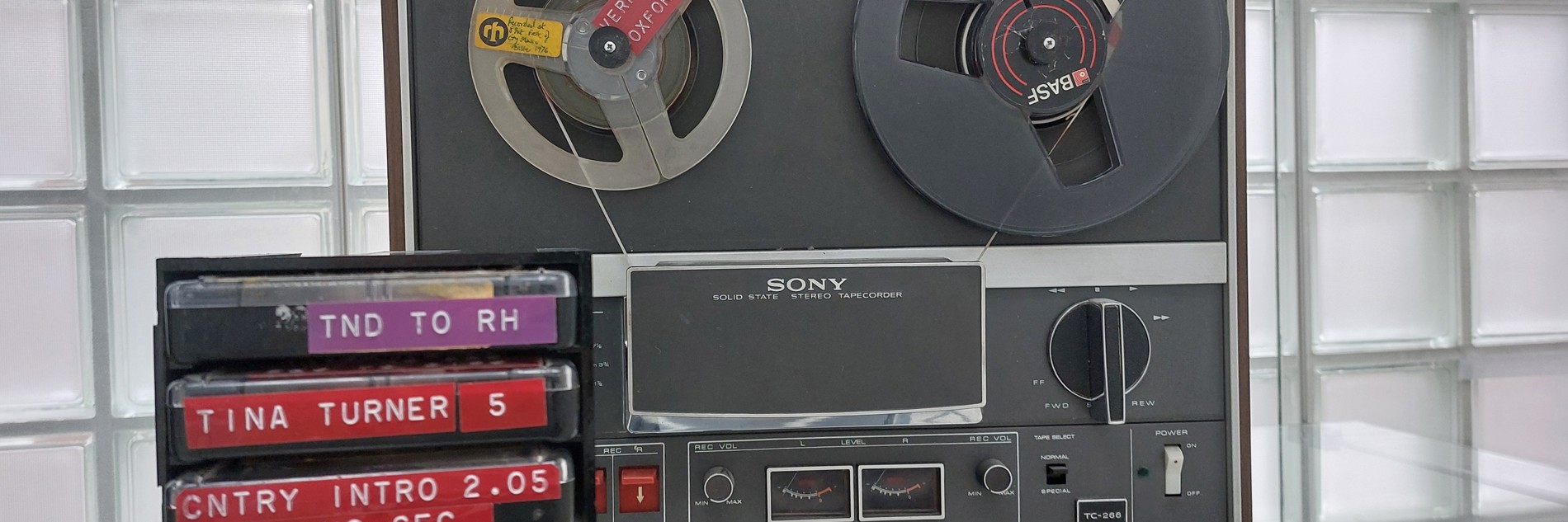 A photograph of radio equipment on display in Millennium Gallery. There is a large machine with two reels in, then a stack of cassette tapes. They are labelled from top to bottom, as follows: TND TO RH, TINA TURNER 5, CNTRY INTRO 2.05 NO SEC, ELVIS PRESLEY NO SEC, and FLETCHERS BREAD