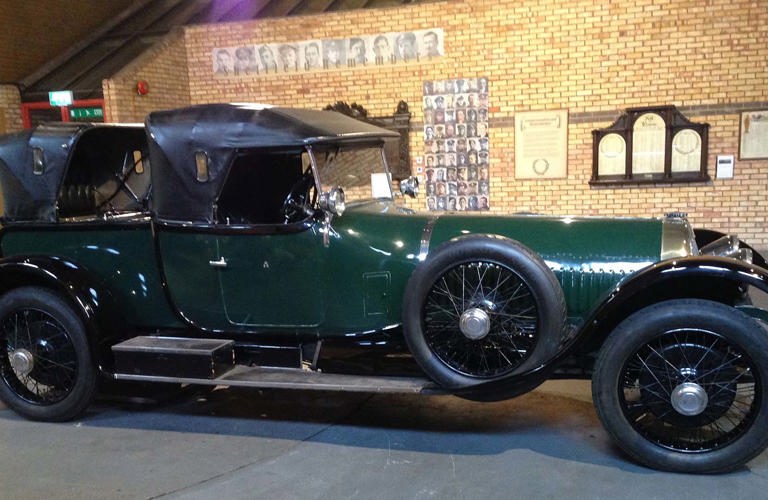 Side elevation of a large vintage car in dark green with a black leather convertible roof, chromed lamps and spoked wheels.