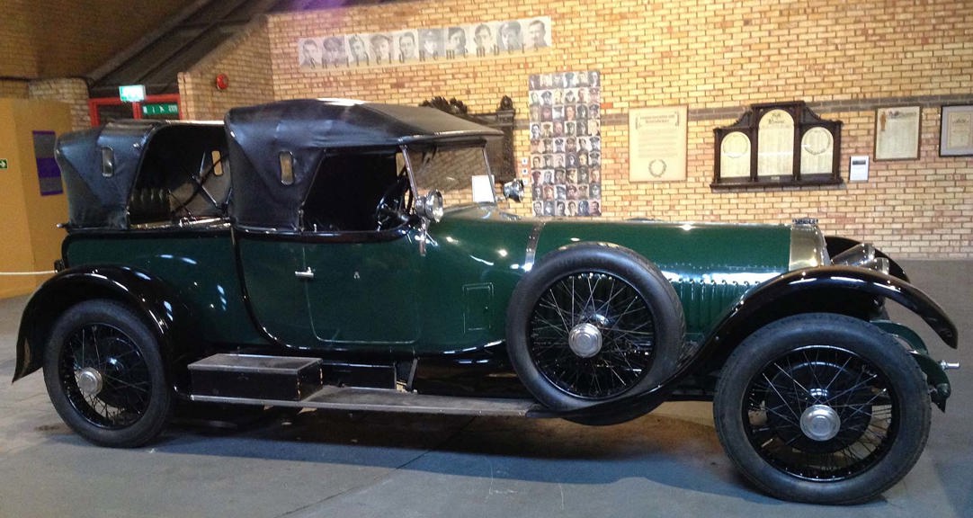 Side elevation of a large vintage car in dark green with a black leather convertible roof, chromed lamps and spoked wheels.